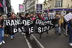 Cortejo en una manifestación con carteles y una pancarta que evoca la lucha trans y contra el SIDA.