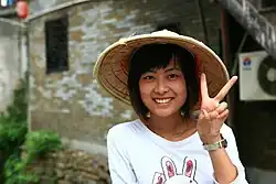 Una mujer china con cabello corto negro, sombrero chino y camiseta blanca posando frente de una edificación, mirando hacia la cámara, sonriendo y haciendo un símbolo de paz con la mano.