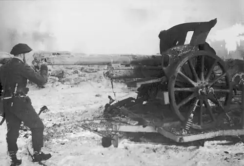 Fotografía en blanco y negro de Churchill uniformado mirando a través del cañón de un gran arma de artillería con una barricada de piedra visible detrás
