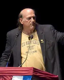 Balding man with long hair, T-shirt and jacket at podium.