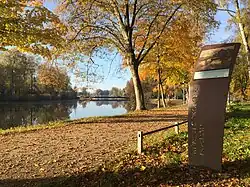 Stele auf dem Künstlerweg Dachau mit einem Bild von Ludwig Dill, neben dem Weg fließt die Amper. Es ist Herbst.