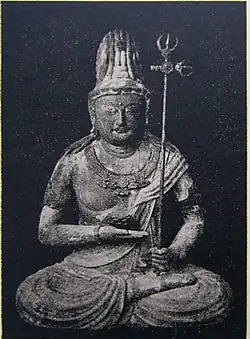 Front view of a statue in lotus position. The palm of the right hand is turned upward and held in front of the stomach. The left hand, close to the right foot, is holding a long pole with decorations at the end resembling a trident.
