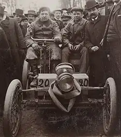 Photo de face de deux hommes assis dans une voiture rudimentaire et portant le numéro de course 20.