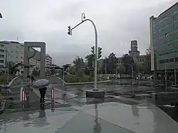 La imagen muestra La Plaza de América tras la remodelación con la Puerta del Atlántico enfrente mientras llueve y un transeúnte espera a cruzar la calle.