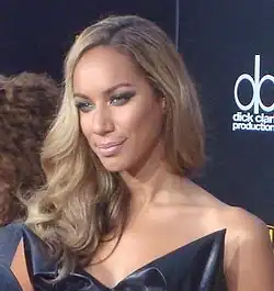 Una mujer con cabellera rubia sonríe mientras se encuentra de pie en una alfombra roja. Ella viste un vestido negro.