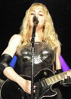 A woman in black clothing holding a guitar and standing behind a microphone stand with one arm extended straight into the air. In the background is a screen with shades of pink and purple.
