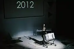 Una persona tocando un tambor, delante de una pantalla que pone 212