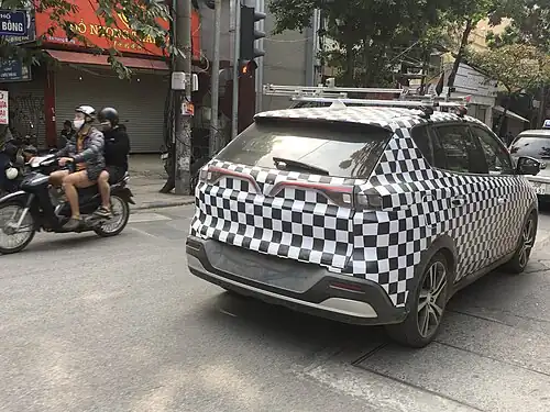 Se observa un auto eléctrico, una SUV modelo VinFast VF e34, con ploteado en cuadriculado blanco y negro, estacionada en una calle Hanoi. En la calle circula además una motocicleta con dos personas, en dirección contraria a la del vehículo. Hay árboles y letreros en los alrededores.