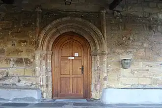 La puerta principal por la que se accede a la Ermita es de medio punto, con tres arquivoltas estriadas con capiteles lisos y flanqueada por robustas columnas también estriadas y rematadas con capiteles idénticos. A la izquierda de la puerta se puede ver la aguabenditera junto con dos bancos corridos de piedra adosados a la pared, en ambos lados.