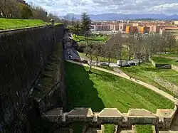 Photo shows an old wall at left and modern buildings at right with mountains in the distance.