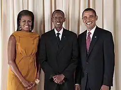 Michelle Obama, Paul Kagame y Barack Obama, de pie y sonriendo frente a una cortina