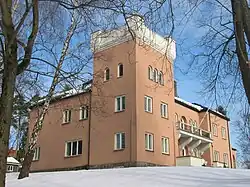 Un edificio importante en piedra rosada o revestimiento, formado por una torre de tres pisos con alas que tienen dos. El edificio se alza, enmarcado por árboles, en un jardín cubierto de nieve.