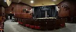 Sala de teatro vacía con butacas. Al fondo un escenario en el que hay una escalera de mano.