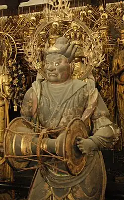 One of the Twenty eight attendants. Three-quarter view of a statue carrying a handdrum in front. A halo is visible around his head.