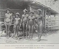 Black-and-white photograph of five nearly naked young women and girls standing in front of a hut; a sixth is visible in the background.