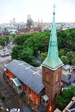 La iglesia vista desde arriba, con su aguja y su techo de teja. El parque colindante lleno de árboles de copa oscura. A la distancia se ve una catedral de piedra arenosa.