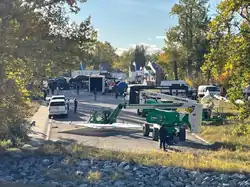 Edificios lejanos rodeados de árboles, con varios vehículos, grúas y personas en primer plano.