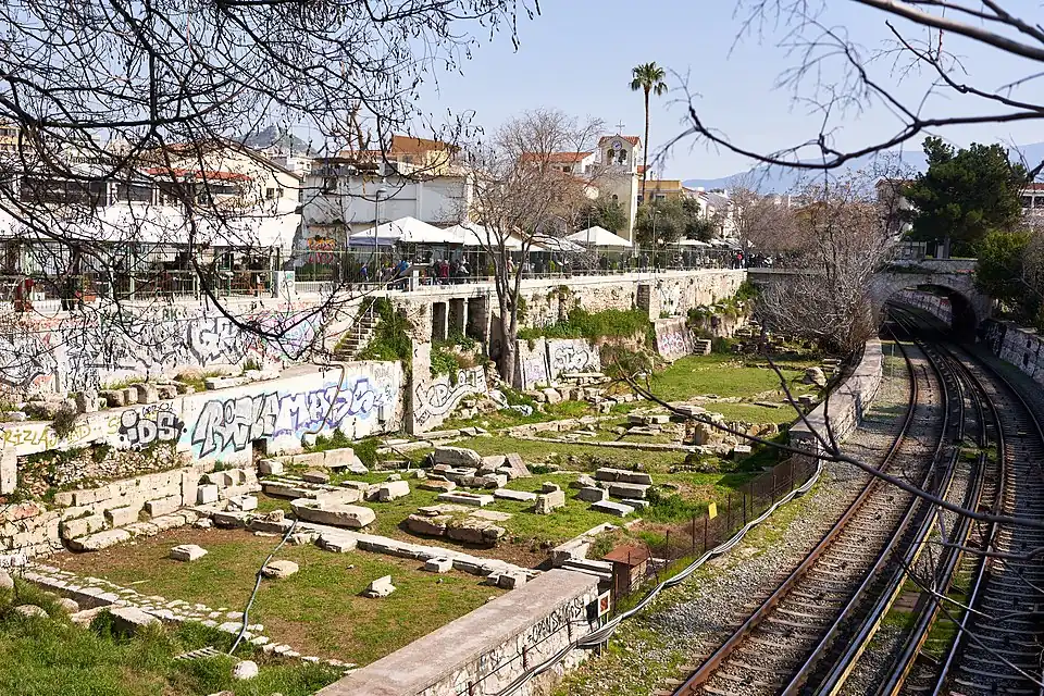 Estoa Basileos desde su pared posterior con las bases de sus columnas internas. El «Desagüe grande» pasaba por su frente (en segundo plano, arriba y a la derecha de la imagen).