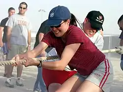 En la imagen se observa en el centro a una mujer de 20 a 30 años, vestida con ropa deportiva, con un short gris con dos rayas rojas en el centro y una remera roja, tomando con sus dos manos una soga grande y tirando la soga hacia el centro de su cuerpo, en el juego de la cinchada. Alrededor de ella, hay dos hombres como espectadores, y detrás de ella, hay otros dos hombres que la ayudan a tirar de la soga.