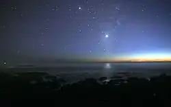 A photograph of the night sky taken from the seashore. A glimmer of sunlight is on the horizon. There are many stars visible. Venus is at the center, much brighter than any of the stars, and its light can be seen reflected in the ocean.