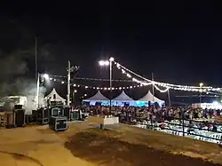 Se aprecia desde arriba en el lado izquierdo cajas del grupo de música, al fondo unas carpas blancas y una gran cantidad de gente bailando. Destacan de la imagen las luces que envuelven la verbena